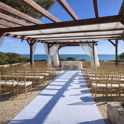 Outdoor wedding setup with chairs and a white aisle runner under a wooden pergola by the sea.