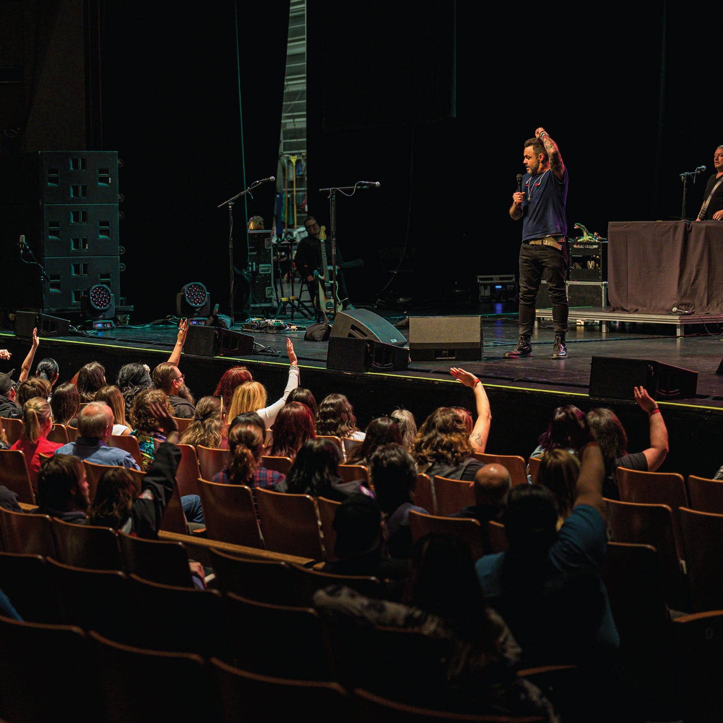 Justin Furstenfeld on stage with raised hand addressing an audience in a theater setting for Blue October's VIP Meet & Greet
