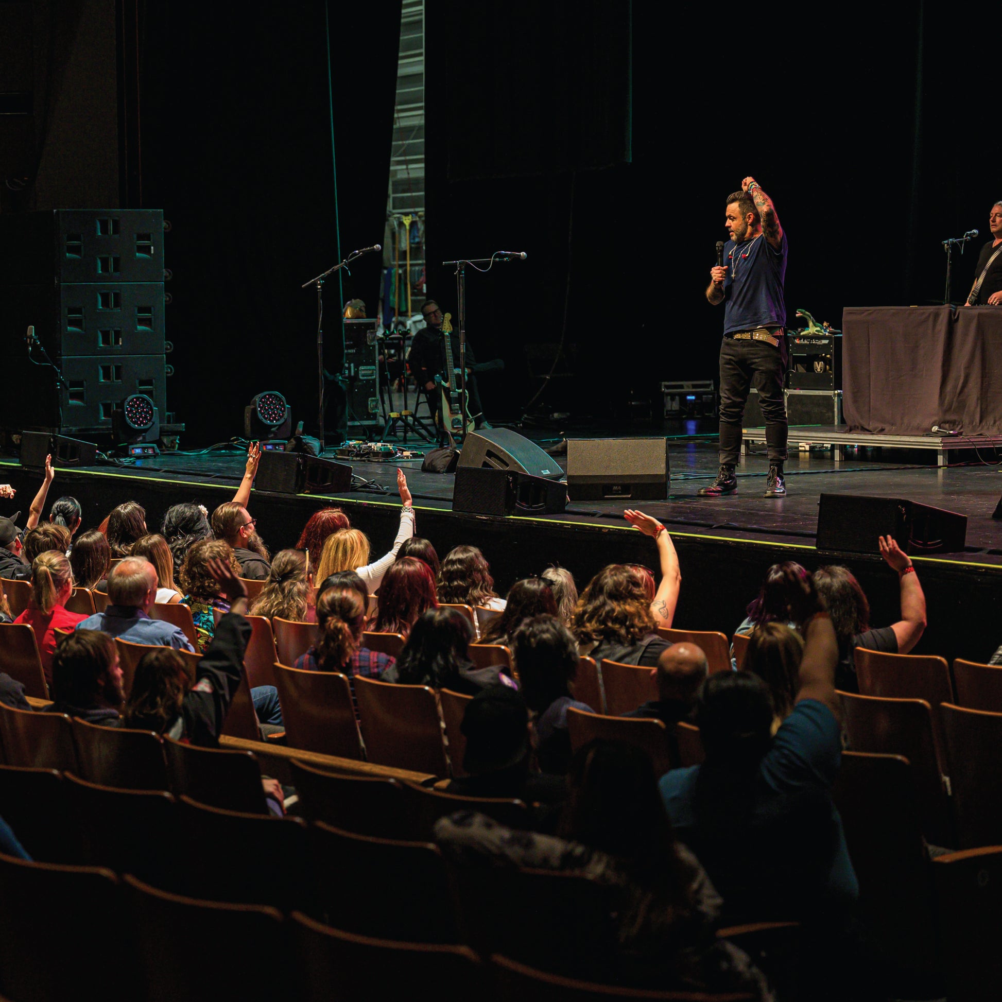 Justin Furstenfeld on stage with raised hand addressing an audience in a theater setting for Blue October's VIP Meet & Greet